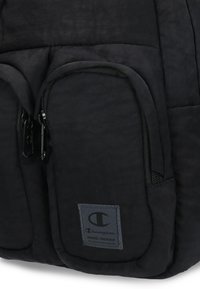 Mochila de tela negra con múltiples bolsillos con cremallera, una superficie texturizada y un parche del logotipo de Champion destacado.