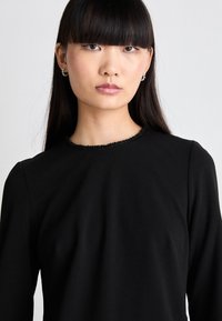 Must mustärg pikkade varrukatega pluus ümmarguse kaelusega, millel on tekstuurne must bead aktsent kaeluse ääres. Sile kangas ja keha lähedane disain.