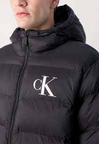 Schwarze Daunenjacke mit durchgehendem Reißverschluss und Kapuze. Verfügt über ein weißes "cK"-Logo auf der Brust und hat eine glatte, glänzende Stoffstruktur.