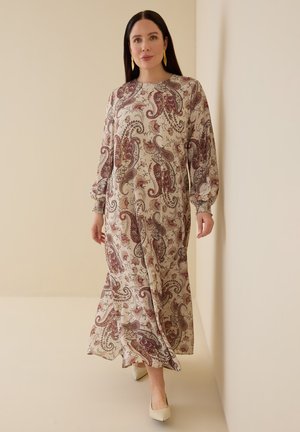 Kvinde med langt mørkt hår iført en beige maxikjole med paisley-mønster og cremede hæle, stående indendørs.