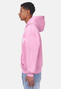 Rosa Kapuzenpullover mit weißem Text, weicher Stoff, Kängurutasche, gerippte Bündchen und Saum, Modell seitlich dargestellt.