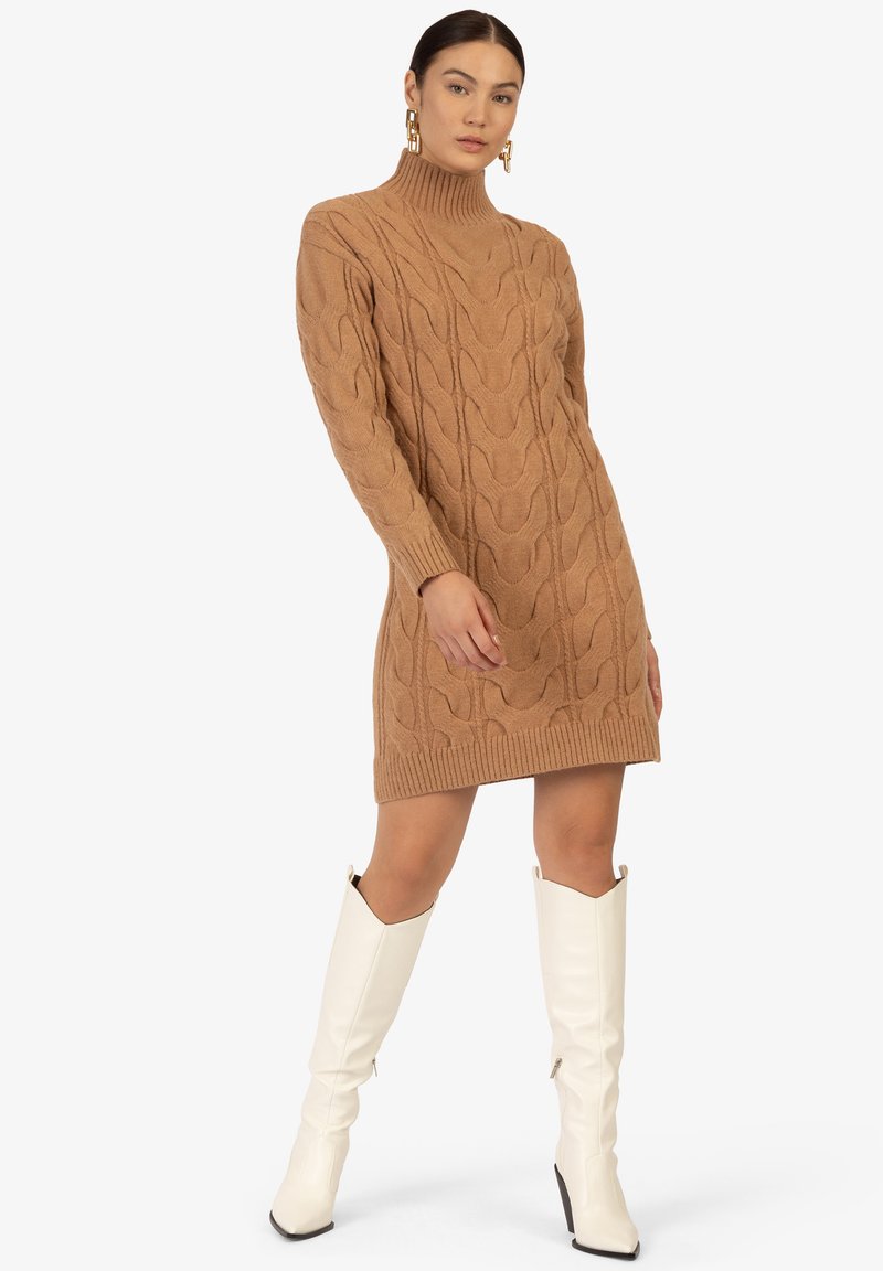 Kamel-gefärbtes Strickkleid mit hohem Kragen, ausgestattet mit einem strukturierten Zopfmuster und gerippten Bündchen, kombiniert mit weißen kniehohen Stiefeln.