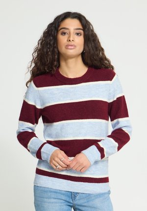 Femme aux longs cheveux bouclés portant un pull rayé bordeaux, bleu clair et blanc et un jean bleu clair, debout, les mains jointes.