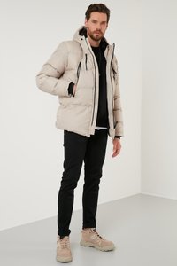 Giacca a vento beige con cappuccio, dotata di dettagli neri sulle zip e tasche, indossata sopra un top nero, abbinata a pantaloni neri e calzature chiare.