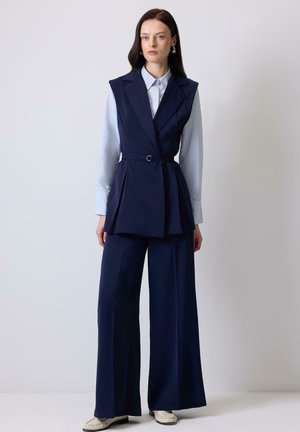 Femeie cu păr închis la culoare, purtând o cămașă albastru deschis sub un vestă fără mâneci, bleumarin, cu curea, și pantaloni largi bleumarin, cu pantofi de culoare deschisă.