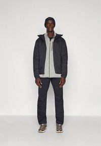 Zwarte geïsoleerde jas over een grijze fleece met ritssluiting, gecombineerd met een zwarte broek en bruine wandelschoenen, met een zwarte muts op het hoofd.