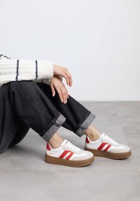Zapatillas blancas con rayas rojas, con una suela tipo plataforma y un upper texturizado. Llevadas con pantalones de mezclilla oscuros arremangados.