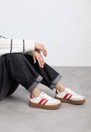 Zapatillas blancas con rayas rojas, con una suela tipo plataforma y un upper texturizado. Llevadas con pantalones de mezclilla oscuros arremangados.