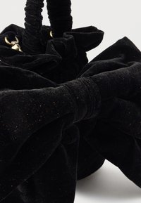 Μαύρη βελούδινη τσάντα χειρός με πτυχώσεις και μεγάλο φιόγκο ως διακοσμητικό. Το υλικό είναι απαλό με λεπτές χρυσές νιφάδες, προσφέροντας μια πολυτελή εμφάνιση.