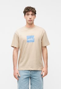 Νέος άνδρας που φοράει μπεζ μπλουζάκι με μπλε τετράγωνο και λευκό λογότυπο σε στυλ γραφής, συνδυασμένο με ανοι χτόχρωμο μπλε τζιν, στέκεται μπροστά σε απλό λευκό φόντο.