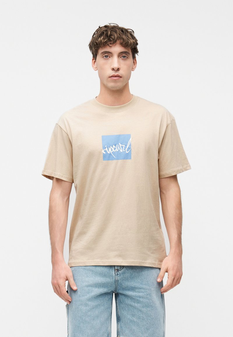 Νέος άνδρας που φοράει μπεζ μπλουζάκι με μπλε τετράγωνο και λευκό λογότυπο σε στυλ γραφής, συνδυασμένο με ανοιχτόχρωμο μπλε τζιν, στέκεται μπροστά σε απλό λευκό φόντο.