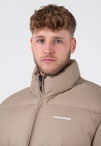 Braune Puffjacke mit hohem Kragen, ausgestattet mit einem Frontreißverschluss und einem weißen "PEGADOR"-Logo auf der Brust. Glatte, gepolsterte Textur.