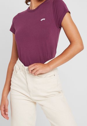 Vrouw draagt een aansluitend paars T-shirt met korte mouwen en een klein wit skateboardlogo, gecombineerd met een hoogwaardige, off-white broek.