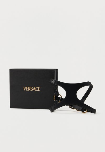 Μαύρο δερμάτινο λουρί σκύλου με εξαρτήματα σε χρυσή απόχρωση δίπλα σε ένα μαύρο κουτί με ανάγλυφη τη λέξη «VERSACE» με χρυσά γράμματα.