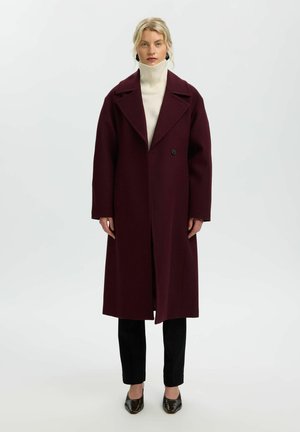 Femme portant un long manteau bordeaux sur un pull à col roulé blanc, pantalon noir et chaussures noires pointues, debout devant un fond uni.