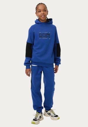 Een blauwe hoodie met contrasterende zwarte mouwen en grafische tekst "FUTURE READY!" in het midden, gecombineerd met bijpassende blauwe sweatpants en multi-kleurige sneakers.