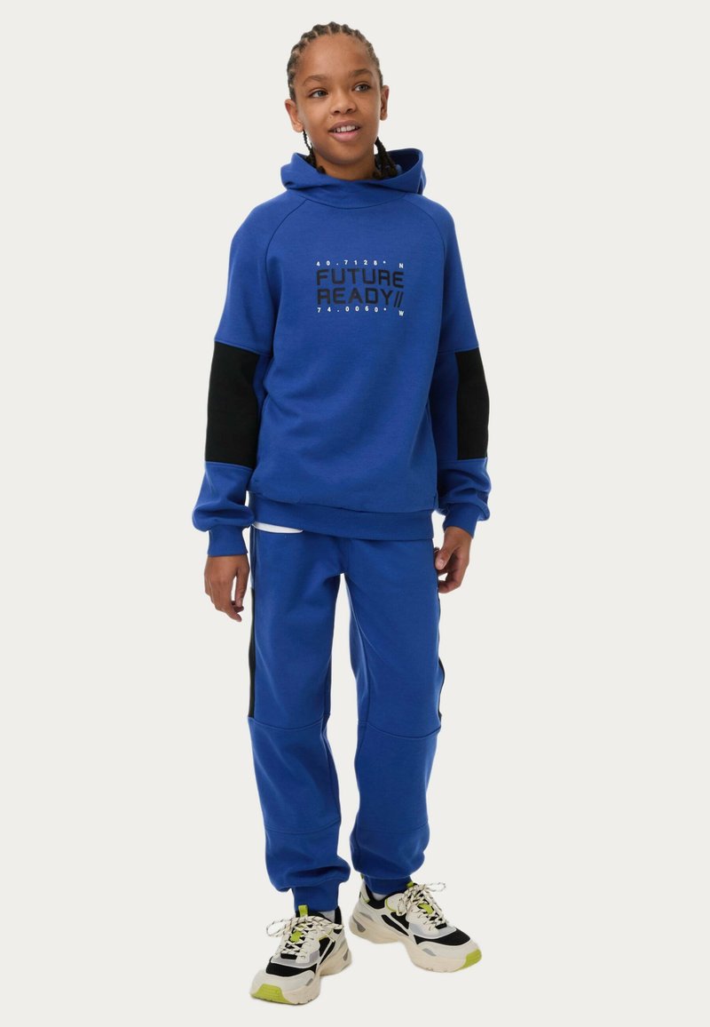 Blå hoodie med kontrasterande svarta ärmar och grafisk text "FUTURE READY!" i mitten, ihop med matchande blå sweatpants och flerfärgade sneakers.