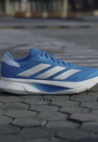 Chaussure de course bleue avec des rayures blanches et une semelle épaisse et amortissante reposant sur une surface pavée en pierre en extérieur.