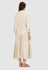 Beżowa, długa sukienka z długim rękawem, z paskiem w talii, warstwową spódnicą i kołnierzykiem zapinanym na guziki. Materiał jest lekki, o gładkiej teksturze i prostym wzornictwie.