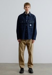 Πουκάμισο από τζιν σε σκούρο μπλε χρώμα με δύο τσέπες στο στήθος, κλείσιμο με κουμπιά και αντίθετη ραφή· συνδυασμένο με μπεζ παντελόνι και μαύρα παπούτσια.