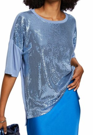 Mujer con una blusa de mangas cortas de color azul claro con brillantes lentejuelas, combinada con una falda azul brillante, sosteniendo un bolso de mano azul oscuro.