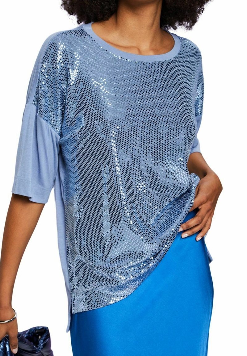 Mujer con una blusa de mangas cortas de color azul claro con brillantes lentejuelas, combinada con una falda azul brillante, sosteniendo un bolso de mano azul oscuro.