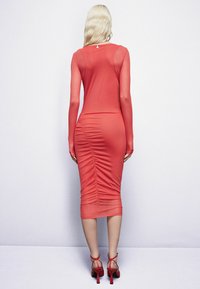 Vestido midi rojo ajustado con mangas largas, detalles fruncidos en la parte posterior y un escote redondo. Combinado con tacones rojos de tiras.