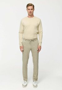 Bézs hosszú ujjú ing világosszürke, texturált, rugalmas derekú nadrággal párosítva. A modellen fehér, szürke részletekkel ellátott sportcipők vannak.