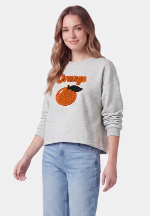 Ung kvinde med langt bølget hår iført en lysegrå sweatshirt med en orange grafik og lyseblå jeans, smilende med en neutral baggrund.