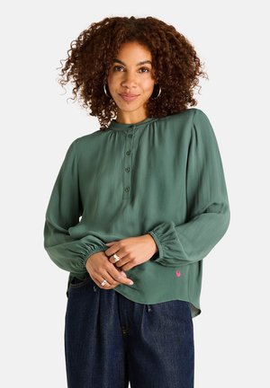 Frau mit lockigem Haar, die eine grüne, langärmlige, geknöpfte Bluse und dunkelblaue Jeans trägt, stehend vor einem schlichten Hintergrund.