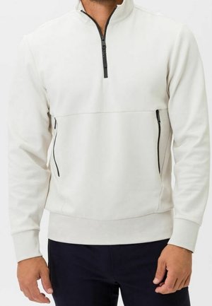 Hombre vistiendo un pulóver blanco con cremallera corta y cremalleras negras en el pecho y en las áreas de los bolsillos, combinado con pantalones oscuros.