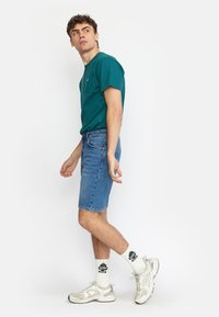 Teal crew neck t-shirt, blauwe denim shorts en zilveren sport sneakers. Witte sokken met zwart grafisch tekst. Casual pasvorm en ontspannen stijl.