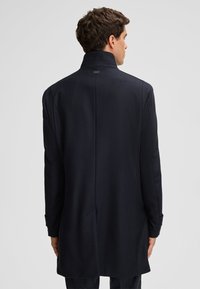 Donker marineblauwe overjas met een hoge kraag, lang ontwerp, gladde textuur, op maat gemaakte pasvorm en minimale brandingdetails op de achterkant.