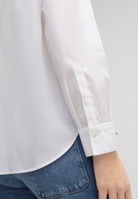 Weißes Hemd mit glatter Textur, ausgestattet mit einem Knopfmanschette und einem abgerundeten Saum. Kombiniert mit blauen Jeans im Denim-Stil mit einer auffälligen Taschendetail.