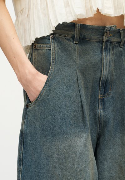 Το χέρι μιας γυναίκας στην μπροστινή τσέπη ψηλόμεσων φαρδιών τζιν παντελονιών, συνδυασμένα με λευκό κροπ τοπ με πιέτες.