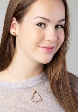 Collier en forme de cœur doré sur une fine chaîne, porté sur un pull texturé gris clair à pois. Des boucles d'oreilles rondes en or complètent le look.