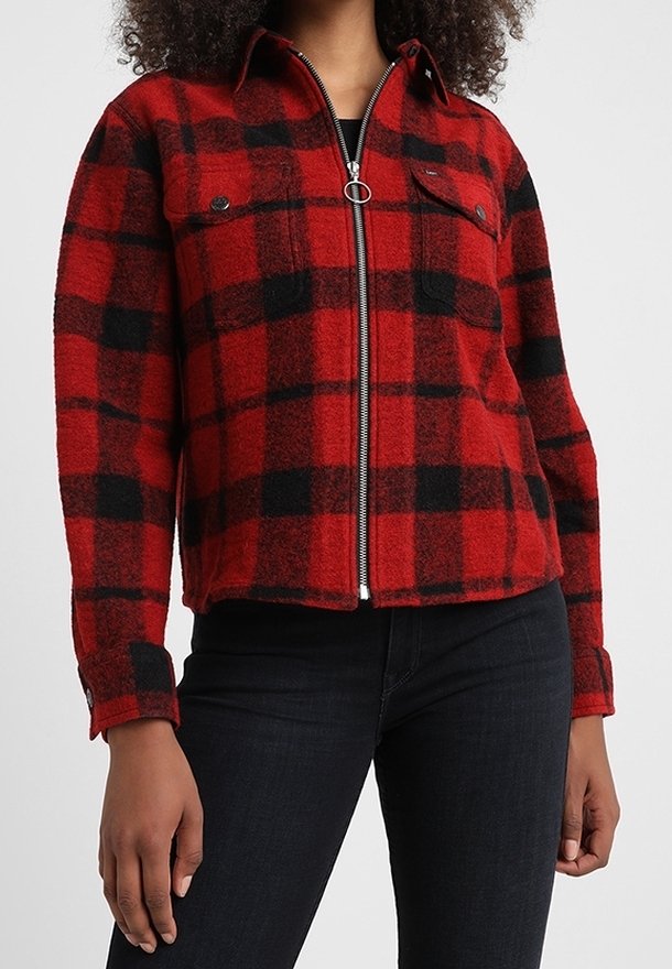 Femme portant une veste zippée à carreaux rouges et noirs avec poches à rabat, associée à un pantalon noir ajusté.