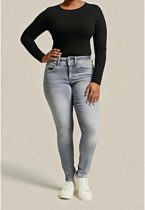 Vrouw draagt een zwart shirt met lange mouwen, lichtgrijze skinny jeans en witte sneakers, staand met één hand op haar heup en de andere ontspannen langs haar lichaam.