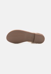 Sole della sandalo piatta realizzata in gomma marrone con scanalature per una migliore trazione. Presenta una parte superiore liscia color beige con un design intrecciato e una fibbia regolabile.