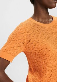 Kortärmad stickad topp i stark orange med ett strukturerat mönster av små hål över hela, med en klassisk rund halsringning.