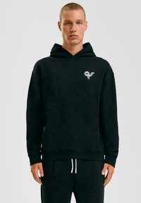 Zwarte hoodie van zacht materiaal, met een voorzak, een hoodie met trekkoord en een klein wit logo aan de linkerkant.