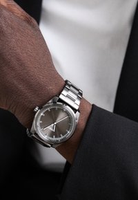 Montre en acier inoxydable argenté avec un cadran gris, présentant un boîtier rond, des aiguilles argentées et un bracelet en métal lisse, portée sur un poignet vêtu d'un costume noir.