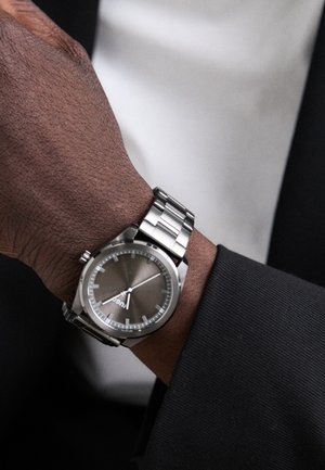 Relógio em aço inoxidável prateado com um mostrador cinza, apresentando uma caixa redonda, ponteiros prateados e uma bracelete de metal lisa, colocado em um pulso com um fato preto.