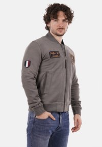 Giubbotto bomber grigio con zip frontale, polsini a coste e due tasche laterali. Presenta toppe sul petto e sul maniche sinistra.