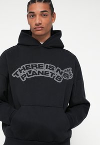 Μαύρο φούτερ από μαλακό ύφασμα, με μεγάλο γκρι γραφικό κείμενο "ΔΕΝ ΥΠΑΡΧΕΙ ΣΧΕΔΙΟ Β" και λεπτομέρειες με φλόγα και σφαίρα. Περιλαμβάνει μπροστινή τσέπη.