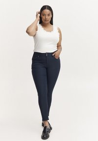 Weißes ärmelloses Top mit gewellten Kanten, kombiniert mit marineblauen Skinny-Jeans mit Fronttaschen und einer Metallknopf-Verschluss. Schwarze Loafers runden den Look ab.