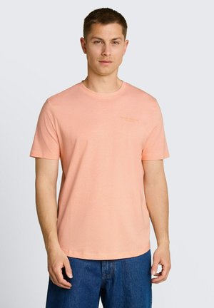 Mężczyzna w jasnopomarańczowej koszulce z krótkim rękawem i niebieskich dżinsach, stoi na tle jednolitego jasnego tła, zwrócony przodem.