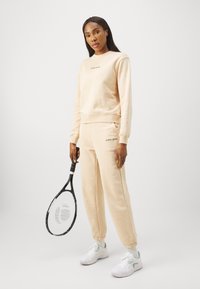 Licht perzikkleurige sweatshirt en bijpassende joggers met elastische boorden. De sweatshirt heeft een geborduurd logo en het model houdt een zwarte tennisracket vast.