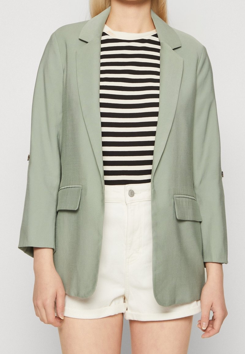 Personne portant un blazer vert clair sur une chemise rayée noire et blanche et un short blanc taille haute, debout devant un fond uni.
