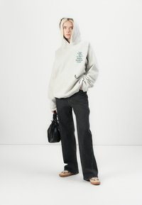 Grey hooded sweatshirt with a green graphic, paired with dark denim jeans and beige slip-on shoes. Holding a small black bag.